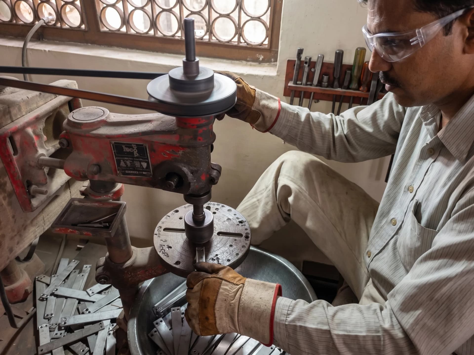 Operator drilling fixing holes into metal hardware plates on a bench drill at the Nexus Fittings factory in Aligarh